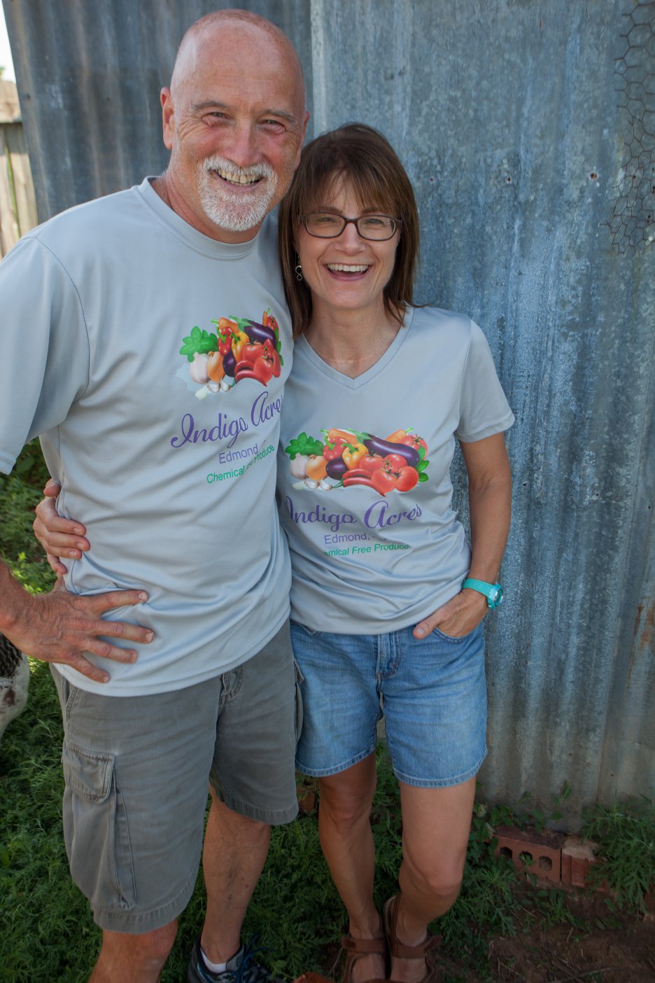 Smiling couple wearing matching gray t-shirts with "Indigo Acres" logo, standing in front of a rustic metal wall, promoting chemical-free produce.