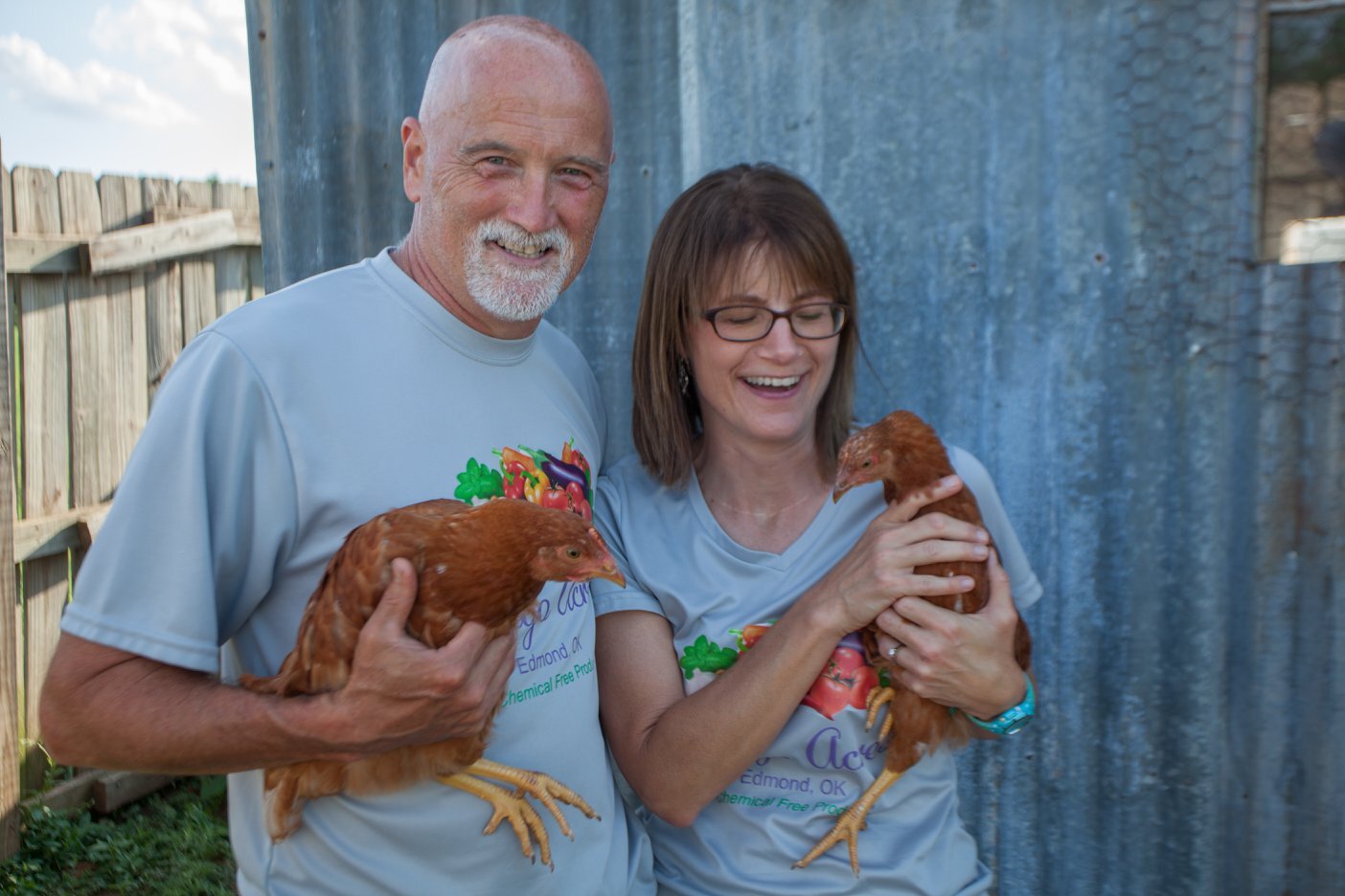 A man and a woman smile while holding chickens in a rural setting, with a rustic wooden fence and a metal shed in the background. They are wearing matching gray shirts promoting a chemical-free farming initiative.
