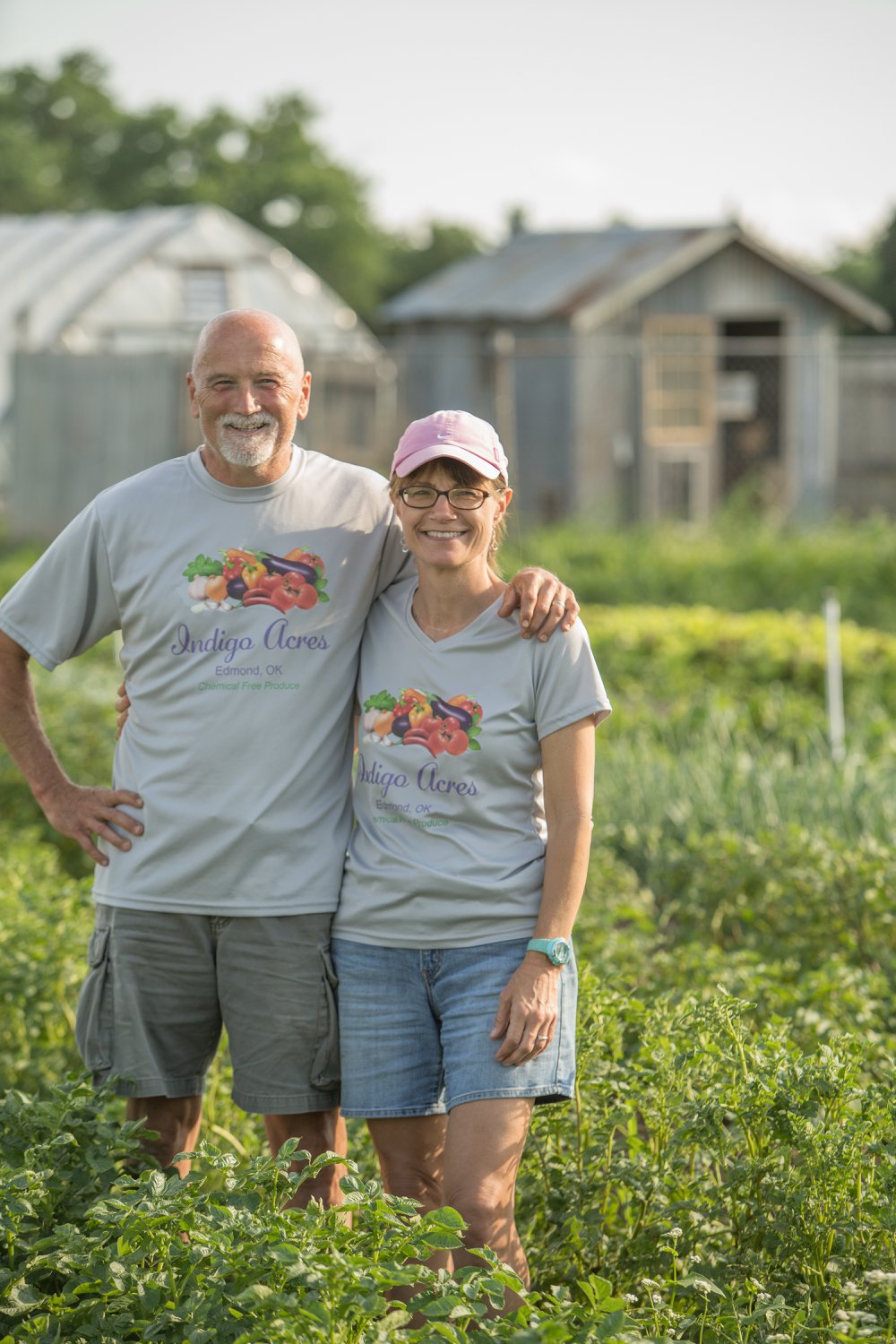 Farmers standing together in a lush green field at Indigo Acres, Edmond, OK, promoting chemical-free produce. The couple is wearing matching gray shirts featuring the farm's logo, surrounded by vibrant crops and greenhouse structures in the background.