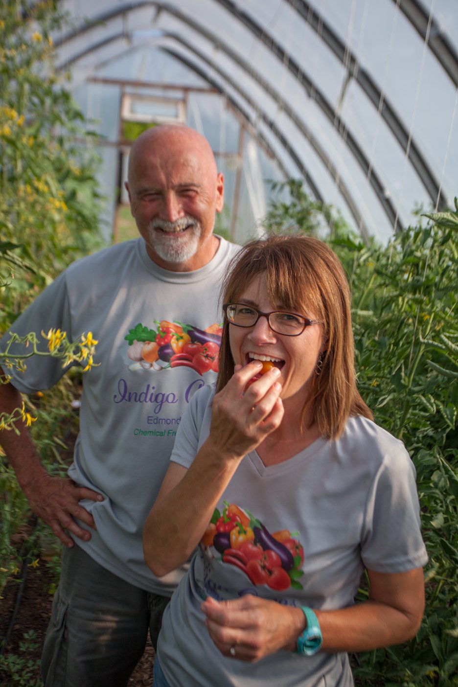 Couple in a greenhouse enjoying fresh vegetables, with the woman tasting a cherry tomato and smiling, while the man stands behind her, both wearing matching shirts featuring colorful produce designs.