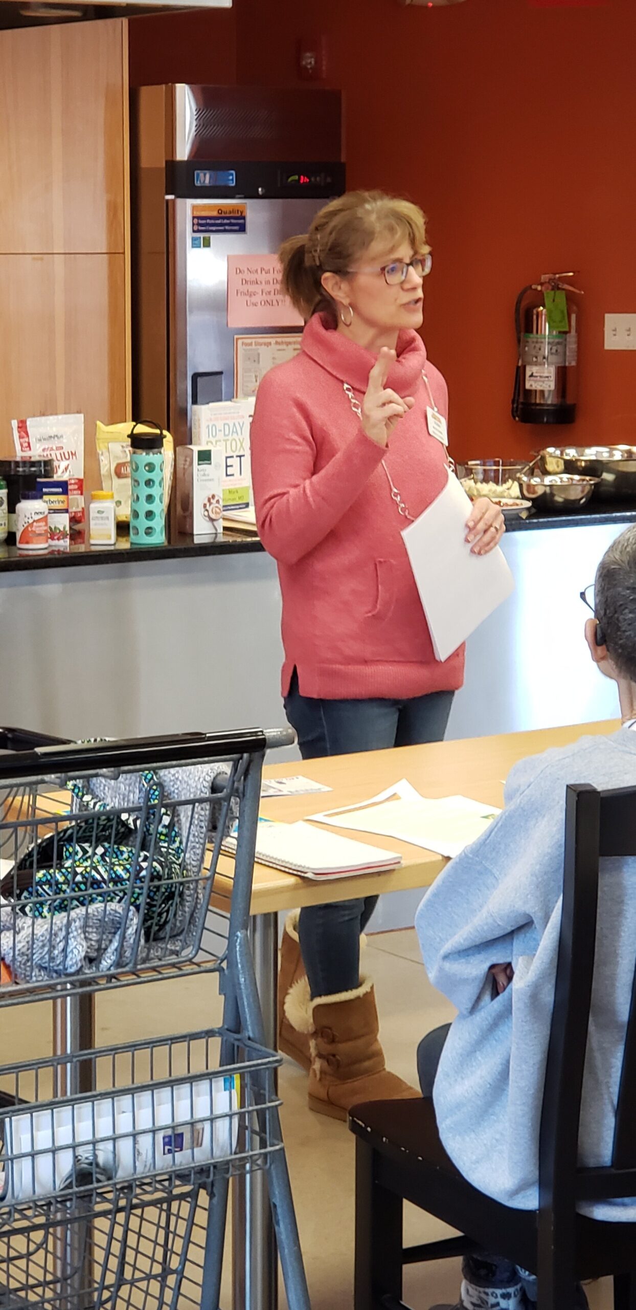A woman in a pink sweater is giving a presentation in a community kitchen setting, surrounded by food and health-related materials. Attendees are seated at a table, engaged in the discussion about nutrition and wellness.
